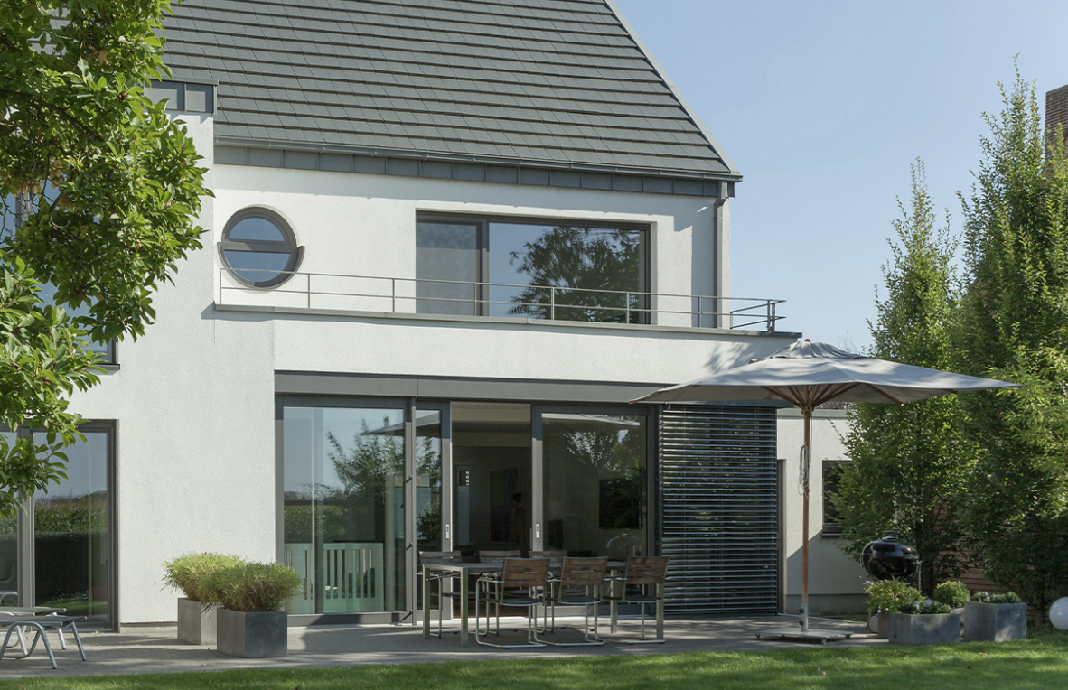 „Modernes Einfamilienhaus mit großer Glas-Schiebetür zur Terrasse, Balkon im Obergeschoss, weißer Fassade und Garten mit Sitzbereich und Sonnenschirm.“